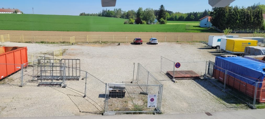 Blick auf einen unbefestigten Platz mit Containern und parkenden Fahrzeugen.